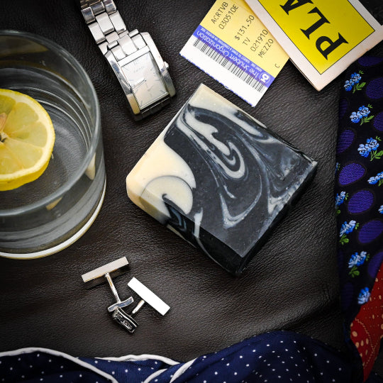 A bar of soap surrounded by cufflinks, a watch, a tie and a glass on a dark background