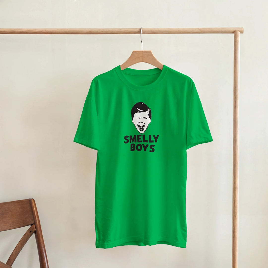 Green t-shirt with 'SMELLY BOYS' text and graphic on a wooden hanger against a beige wall.