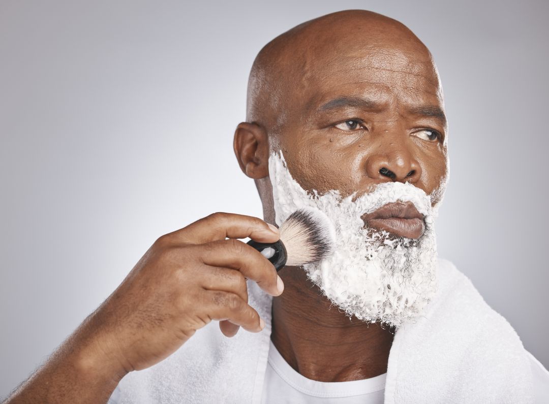 A close-up of a man shaving with white shaving foam on his face, holding a shaving brush against his cheek, with a white towel draped over his shoulders against a light gray background.