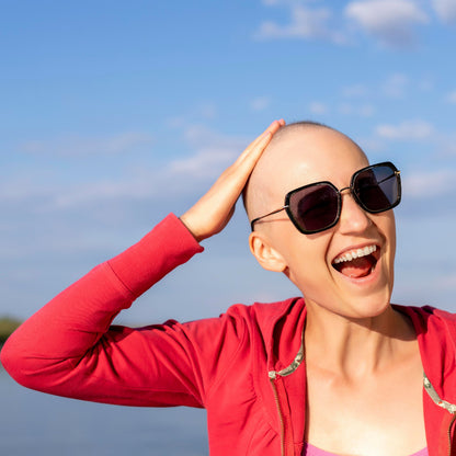 A smiling woman outdoors wearing a red zip-up hoodie, pink top, and black sunglasses against a blue sky background.
