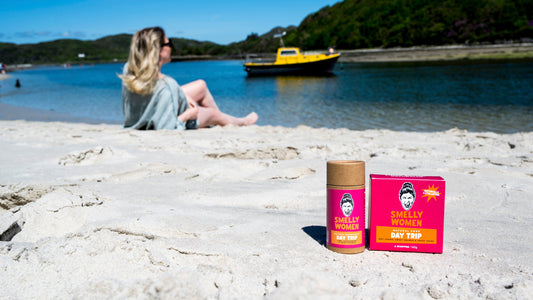 A small tube and matching pink box of Smelly Women Day Trip soap and deodorant are placed on a sandy beach in the foreground, with a woman sitting by the water and a boat blurred in the background.