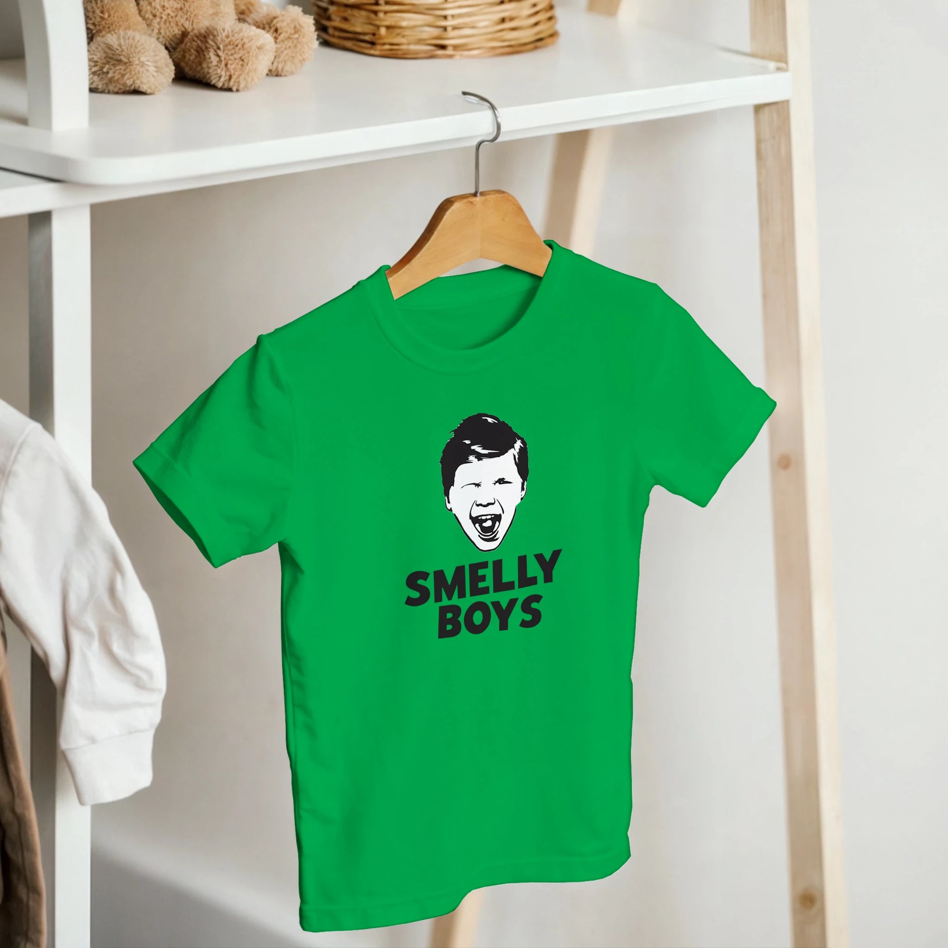 Green t-shirt with 'SMELLY BOYS' text and graphic on a hanger against a white shelf background.