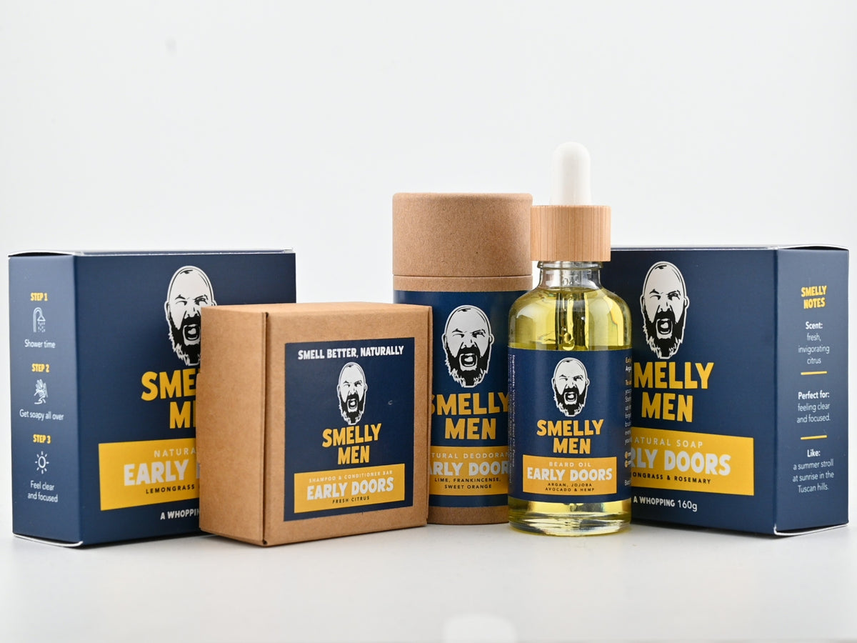 A personal care set for men by Smelly Men, featuring a clear dropper bottle filled with yellow oil, a brown cardboard tube, a brown box, and two dark blue outer boxes with yellow text and a stylized face graphic.