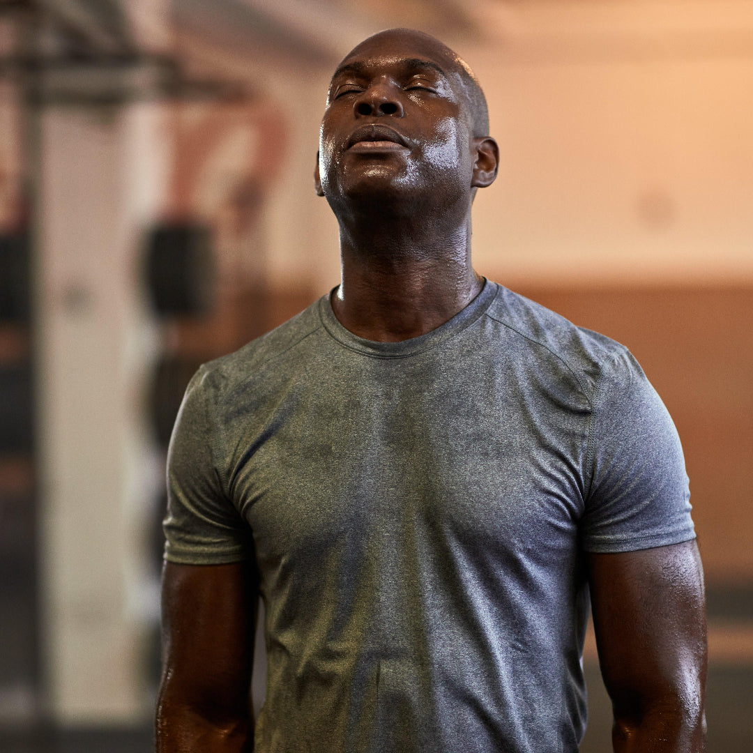 A man in a gym wearing a heather gray athletic t-shirt, captured from the chest up with a blurred warm-toned gym background.