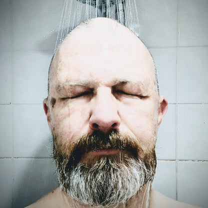 A close-up of a bald man with a full gray and brown beard, standing under a shower head with water running down, against a light gray tiled wall.