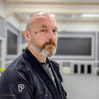 A man with a beard and shaved head wearing a black corduroy martial arts gi jacket with a white 'P' logo on the sleeve, standing in an indoor dojo or training hall.