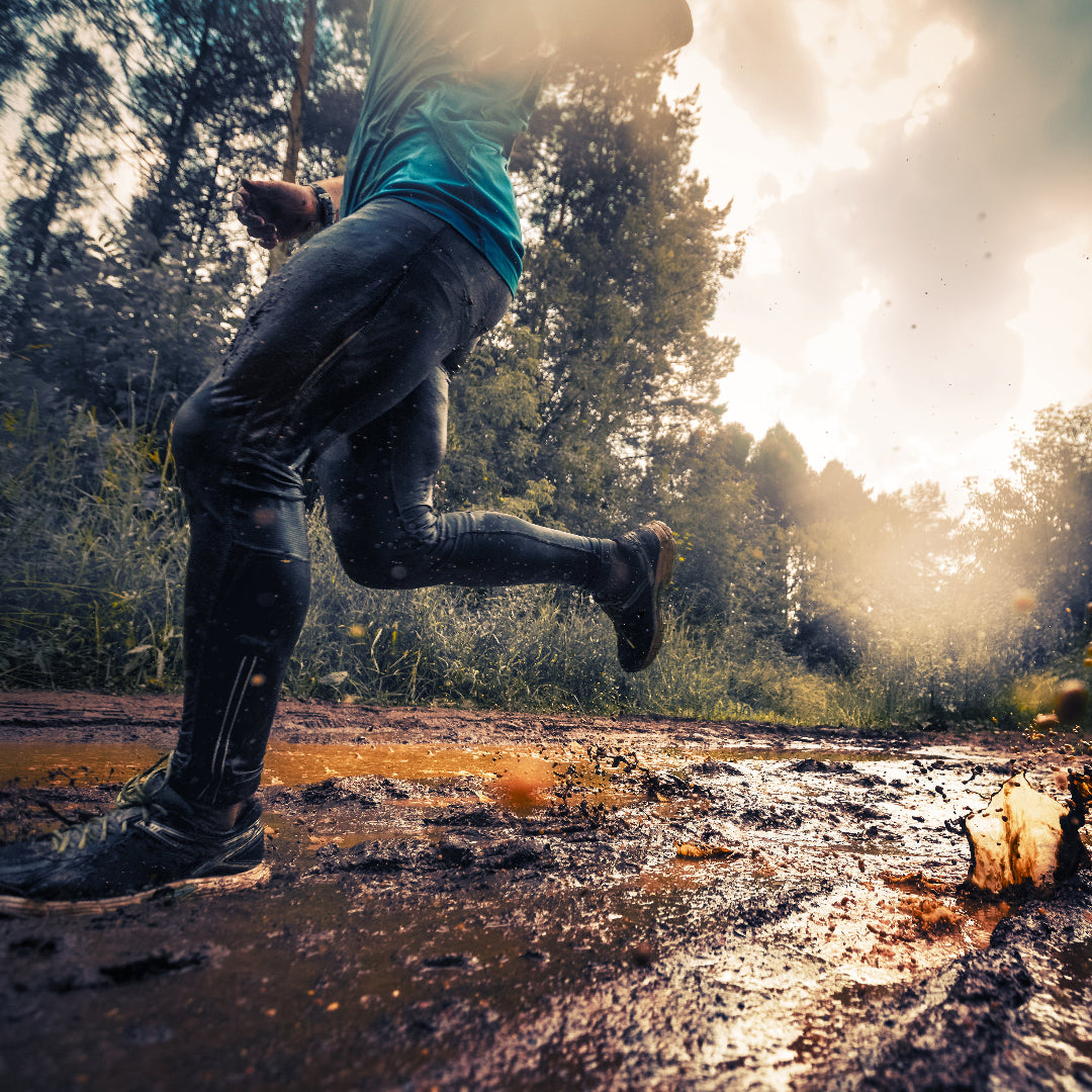 A person running through deep mud in a forest, wearing dark pants, black boots, and a teal top, with dramatic sunlight and clouds in the background.
