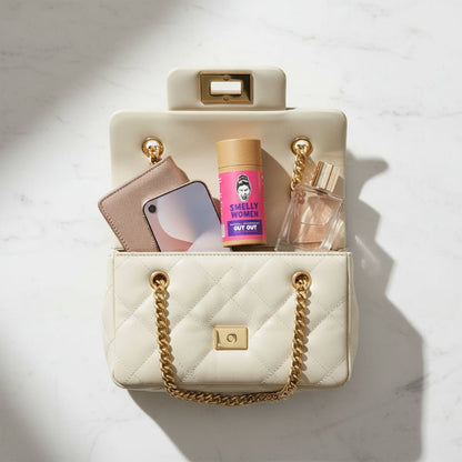 White handbag with gold chain handle containing a phone, wallet, and two bottles on a light background